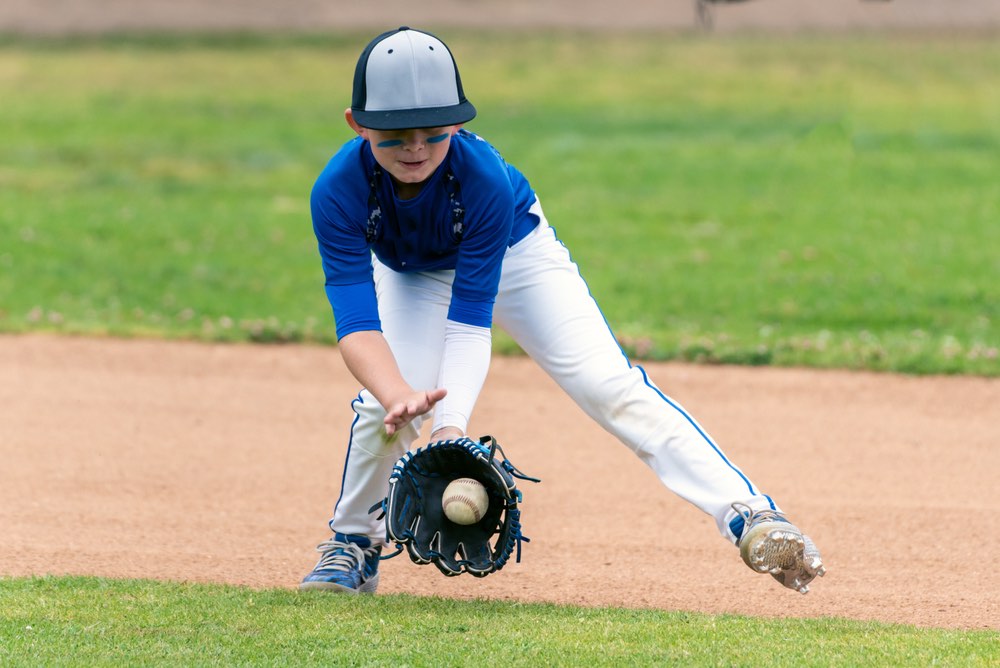 Youth baseball player in blue uniform fielding a ground ball into his glove in the infield during a game.